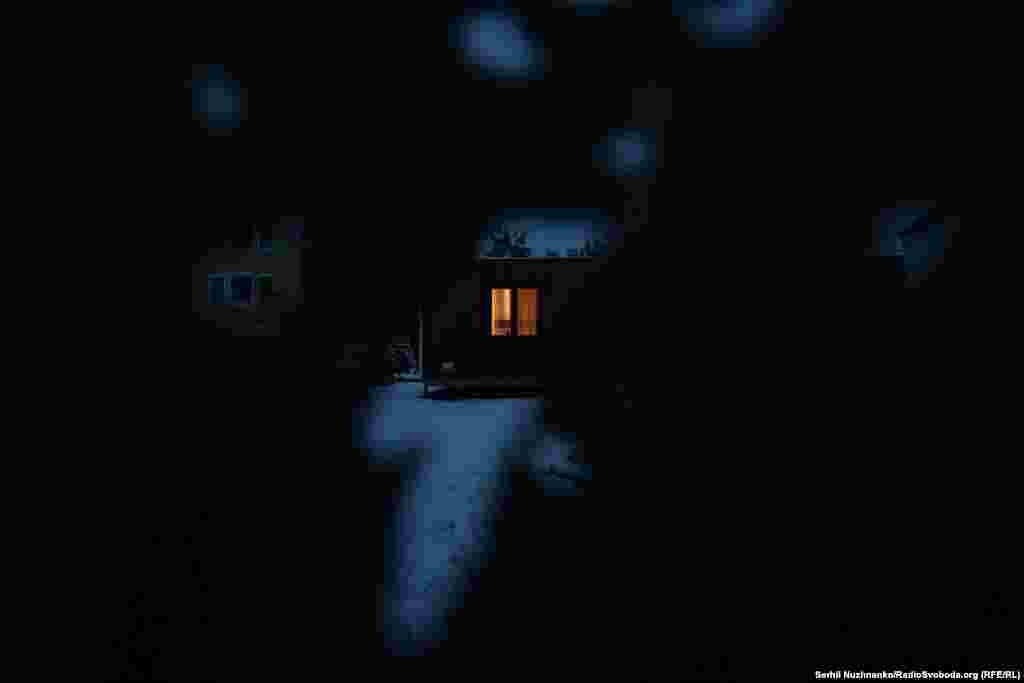 Крізь отвори у посіченому обстрілами паркані видно світло у вікні модульного будиночка і згорілий автомобіль на подвір&#39;ї