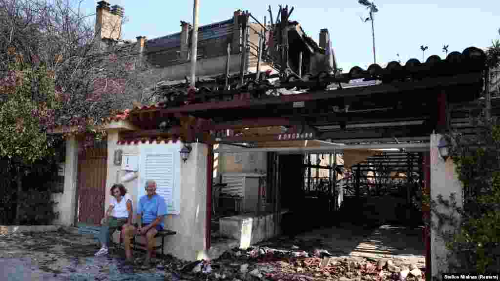 Egy közel kilencvenéves görög házaspár erdőtűz által megrongált otthonuk előtt az Athén melletti Kalíviában július 18-án.&nbsp;Görögországot múlt hét óta hőhullám sújtja, a hőmérséklet az ország középső részén elérte a 44 Celsius-fokot