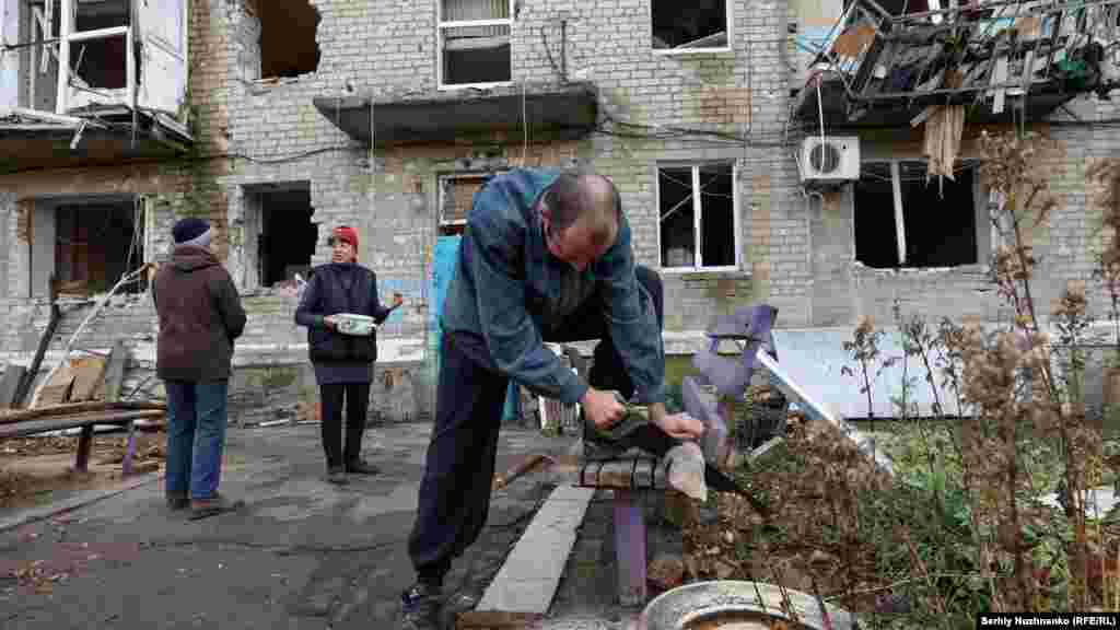 Egy helybeli tűzifát vág, miközben a háttérben a lakhatatlanná vált otthonok előtt beszélget két avgyijivkai.&nbsp;Noha Avgyijivkában van egy humanitárius támogatóközpont, amely élelmet, áramot és menedéket kínál, az emberek számára rendkívül veszélyessé vált odamenni
