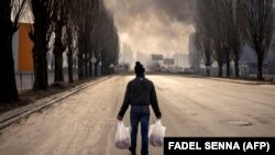 Egy férfi szatyrokat tartva sétál egy égő raktár közelében, amelyet egy orosz gránát talált el Kijev külvárosában 2022. március 24-én. 