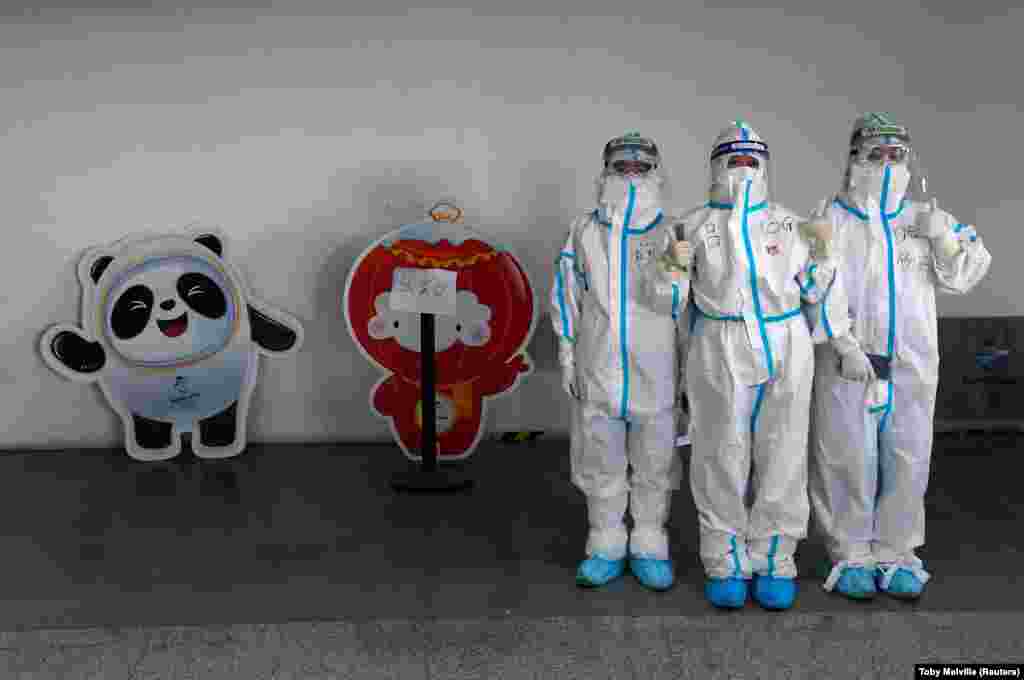 A pekingi téli olimpia kabalafigurája mellett pózolnak védőfelszerelésben a személyzet tagjai a kínai főváros repülőterén január 31-én