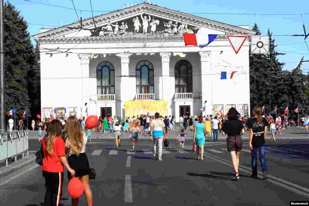 Banorë vendas të Mariupolit duke ecur para Teatrit të Dramës në Donjeck gjatë një festivali me temë franceze më 2019. /&nbsp;Një grua e moshuar kalon pranë teatrit të shkatërruar, ndërsa afër shihet një automjet i blinduar të forcave ruse në prill të këtij viti. &nbsp;