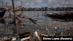 Egy kutya sétálgat és keres élelmet magának a szeméthalmok között, míg a háttérben egy fiú ugrik éppen az erősen szennyezett folyóba.