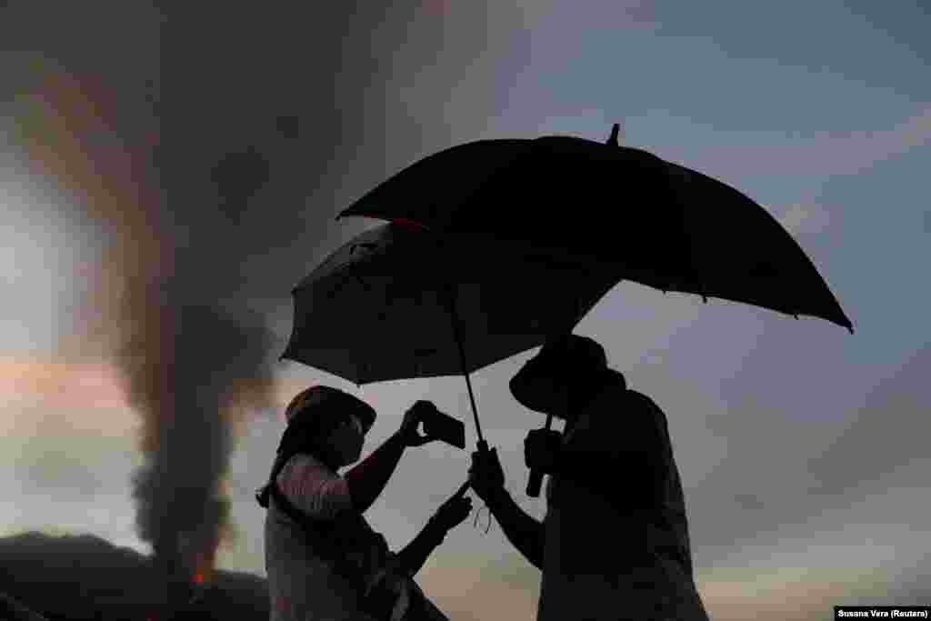 Esernyőt tartanak a fejük fölé a vulkáni hamu miatt, miközben a vulkánnal a háttérben fotózkodnak október 18-án