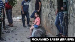 Palesztin lány ül egy fel nem robbant izraeli rakétán az izraeli légi csapások után Gázában, 2021. május 19-én. Május 6-án hat palesztin család erőszakos kilakoltatása miatt a jeruzsálemi Sejk Dzsarrah negyedben összecsapások voltak, amely a negyedik gázai háborúhoz vezetett