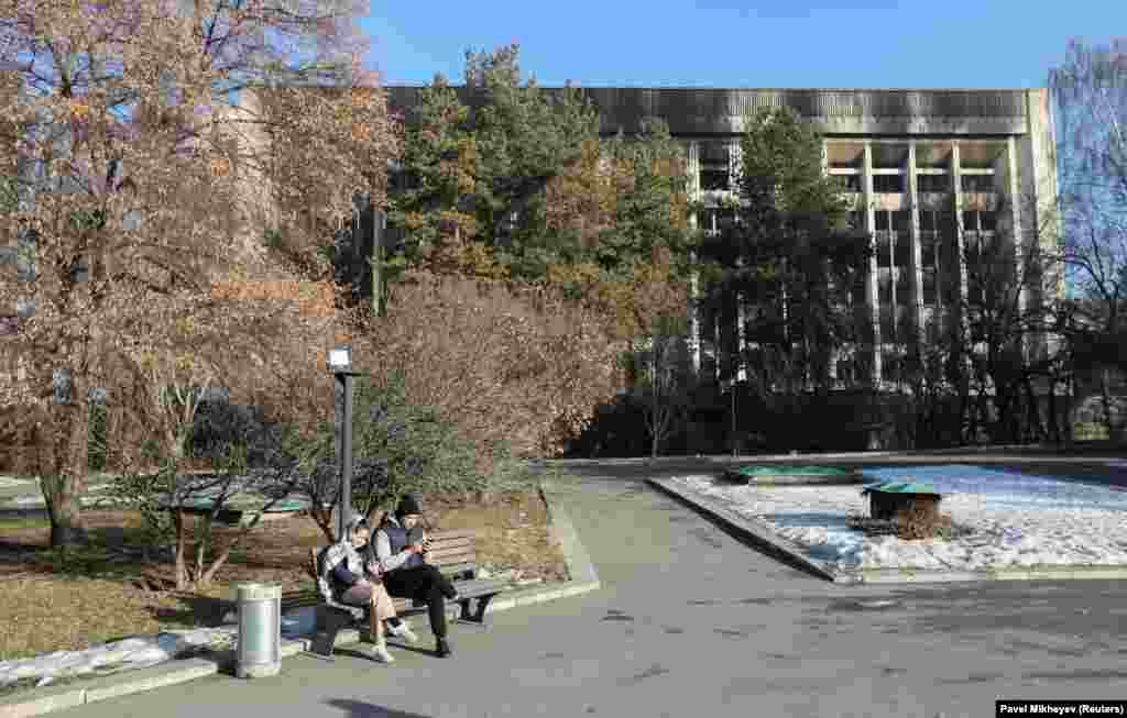 Dy persona qëndrojnë të ulur në një bankë pranë ndërtesës së djegur të Komunës së Almatit më 12 janar.&nbsp; &nbsp;
