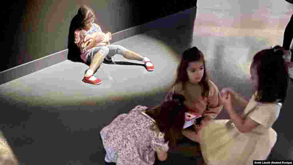 Az előtérben látható három kislány valódi, a fal mellett ülő a szobor. Ha távolabbról nézzük, akkor ez egyáltalán nem egyértelmű. (Particia Piccinini: A gondoskodó)