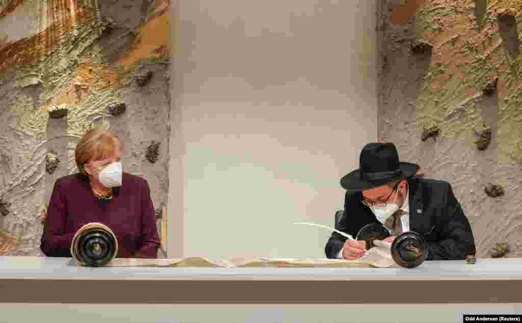 Angela Merkel német kancellár is részt vett a megemlékezésen. Ezen a fényképen azt figyeli, amint Saul Nekrich rabbi beleír a történelmi&nbsp;Sulzbachi Tóratekercsbe. Az idei németországi megemlékezés központi eseménye a sok vihart megélt tóratekercs helyreállításának utolsó mozzanata, a munka lezárása az utolsó betűk szertartásos megírása.