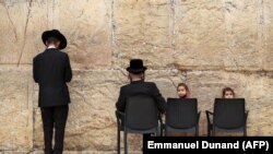 Izraeli gyerekek egy széken ülnek, miközben ultraortodox férfiak a jeruzsálemi Siratófalnál imádkoznak, a fal a judaizmus egyik legszentebb helye.