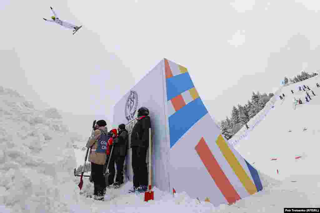 Работники с лопатами в руках наблюдают, как спортсмен совершает прыжок.