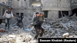 Чоловік несе поранену дитину на тлі зруйнованих будівель в обложеному місті Хамурія, Східна Гута, Сирія, 21 лютого 2018 року