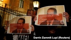 معترضان به کشته شدن خبرنگار مالتی٬ تصویر علی صدر هاشمی‌نژاد را که روی آن نوشته شده «روزنامه‌نگاری جرم نیست. پولشویی جرم است» در دست گرفته‌اند- مالت٬ مارس ۲۰۱۸